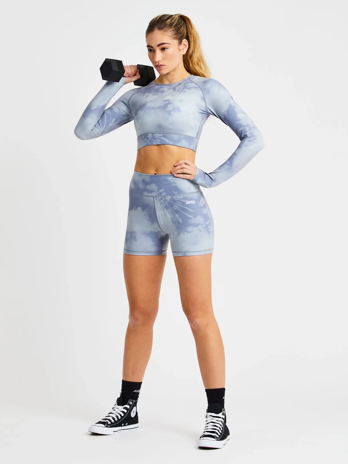 The model is holding a weight and modeling a cropped long sleeve workout top and biker shorts in blue and grey tie dye.