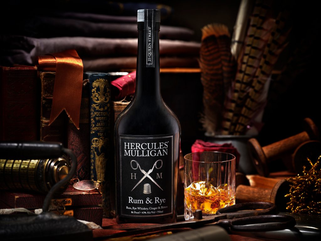 Black bottle of rum with Hercules Mulligan label. Next to it to the right is a glass of rum with ice.